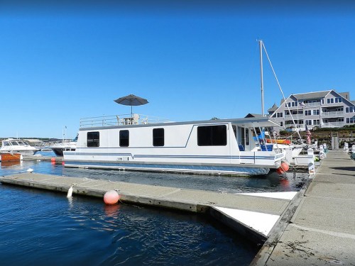 A white houseboat docked at a marina on a clear day, with nearby boats and a building in the background.