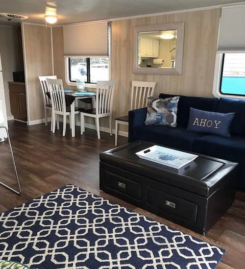 Cozy interior of a trailer with a dining area, sofa, and decorative rug, featuring light wood paneling and bright windows.
