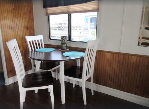 A small dining area with a round table, four chairs, and blue placemats, next to a window with a view of boats.