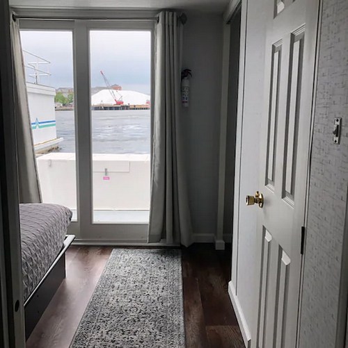 Interior view of a room with a bed, a door, and a large window overlooking water and a distant industrial area.