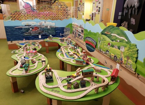 Colorful play area with train tracks, buildings, and landscapes on tables, surrounded by a mural depicting a scenic view.