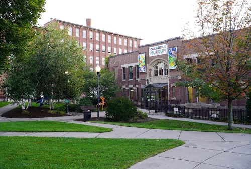 Exterior view of a children's museum surrounded by greenery and pathways, with colorful banners and a brick building.