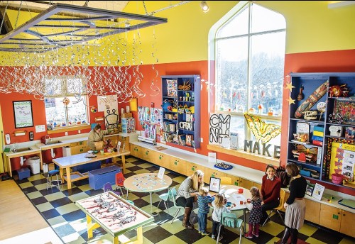 A colorful art classroom with children and adults engaged in creative activities at tables surrounded by art supplies.