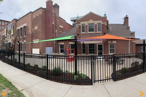 A playground area with colorful canopies, surrounded by a black fence and brick buildings in the background.