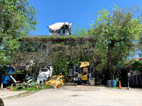 A rundown building covered in vines, with old vehicles and construction equipment scattered in front under a clear blue sky.