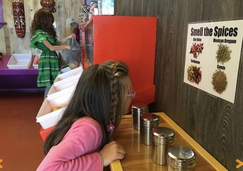 Two children explore a spice display, one smelling spices while the other interacts with a colorful background.