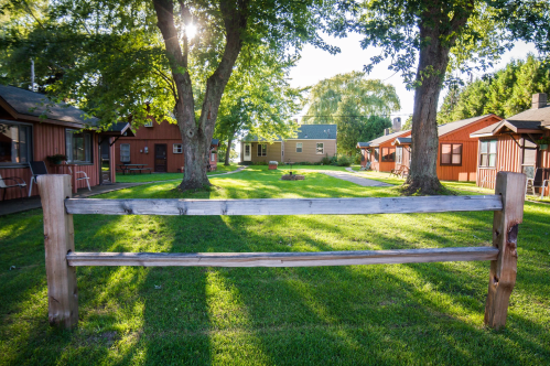A wooden fence in a grassy area with cabins and trees, illuminated by sunlight in a serene outdoor setting.