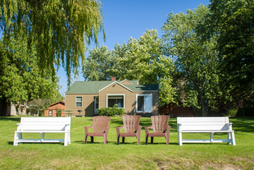 A cozy house surrounded by trees, with four outdoor chairs arranged on a grassy lawn in front.