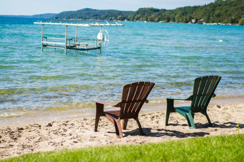 Two Adirondack chairs sit on a sandy beach by a calm lake, with a dock visible in the water. Green hills in the background.