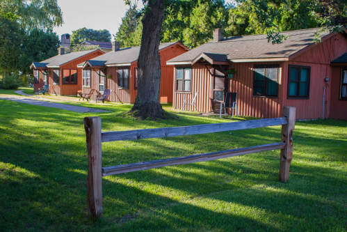A row of rustic cabins with wooden fences, set on a grassy area surrounded by trees.