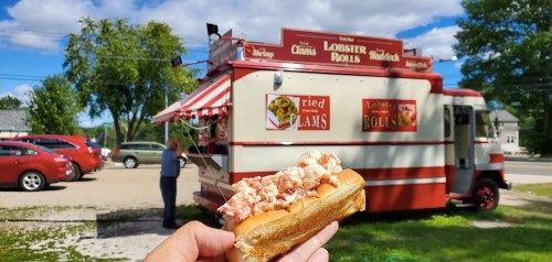 A hand holds a lobster roll in front of a food truck serving lobster rolls and fried clams, with cars parked nearby.