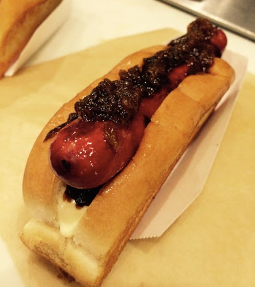 A hot dog in a bun topped with caramelized onions and a drizzle of sauce, served on a paper tray.