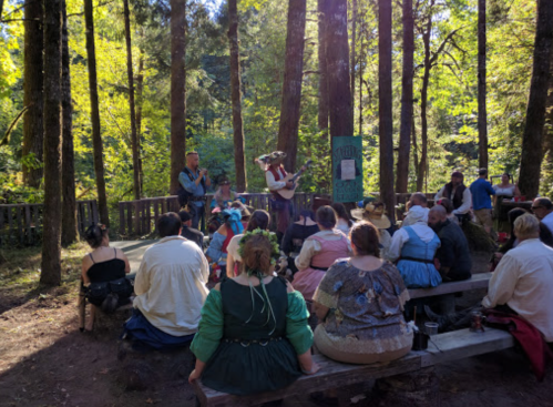 A group of people in period costumes sits on benches in a forest, watching a performance on a wooden stage.