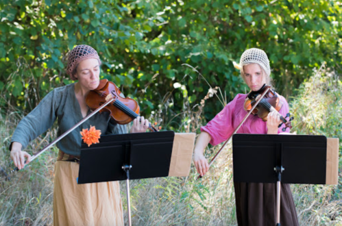 Two women in historical attire play violins outdoors, surrounded by greenery, with music stands in front of them.