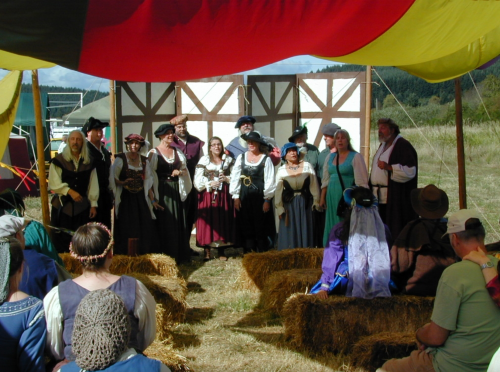 A group of performers in medieval costumes sings on stage under a colorful tent, with an audience seated on hay bales.