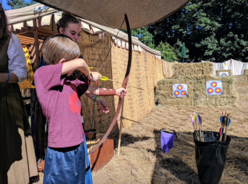 A child aims an arrow at targets while an adult watches, surrounded by hay bales at an outdoor archery event.
