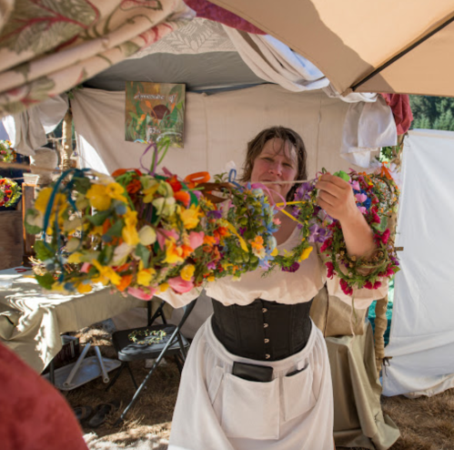 A woman in a white dress holds a large garland of flowers, standing in a tent decorated with colorful fabrics.