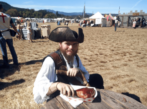 A man in a pirate costume sits at a table, holding a piece of meat, with a festival scene in the background.
