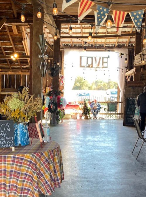 Interior of a rustic barn decorated with lights and banners, featuring a table with flowers and a "LOVE" sign in the background.