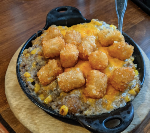A skillet dish with ground beef, corn, melted cheese, and crispy tater tots on top, served on a wooden board.