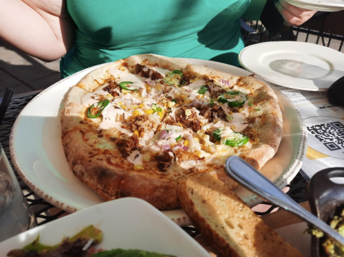 A close-up of a pizza topped with pulled pork, jalapeños, and onions, served on a table with a green shirt in the background.