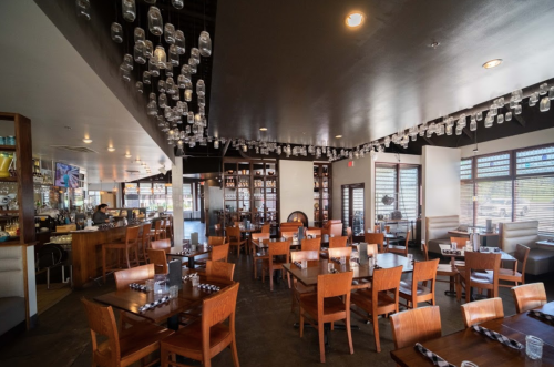 A spacious restaurant interior with wooden tables, chairs, and hanging glass jars, featuring a bar in the background.