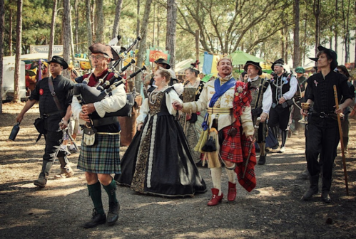 A lively group in historical costumes parades through a forested area, featuring musicians and dancers.