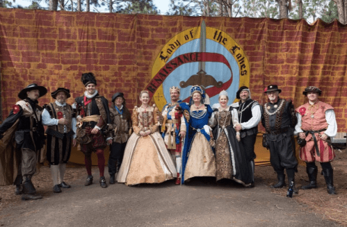A group of people in Renaissance costumes poses in front of a colorful backdrop at a festival.