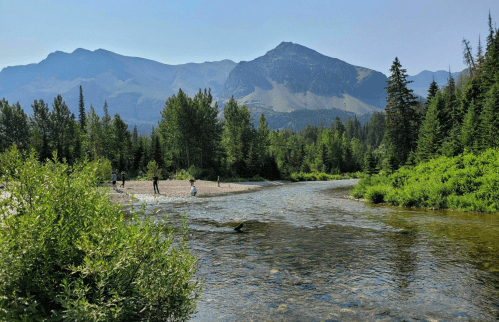 A serene river flows through a lush landscape, with mountains in the background and people enjoying the scenery.