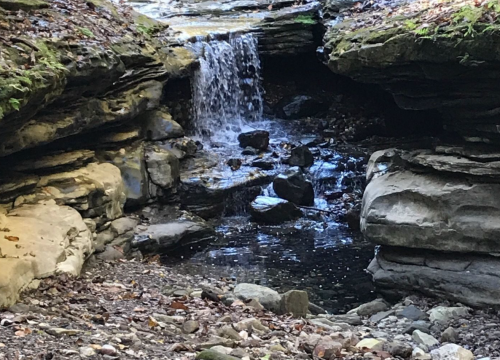 A small waterfall cascades over rocks into a serene pool, surrounded by rugged stone formations and greenery.