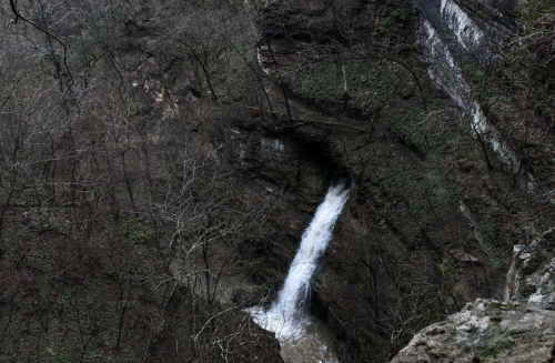 A small waterfall cascades down rocky terrain, surrounded by bare trees and rugged cliffs in a serene, natural setting.
