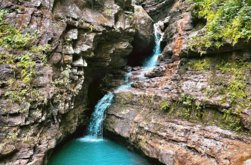 A serene waterfall cascades over rocky cliffs into a turquoise pool surrounded by lush greenery.
