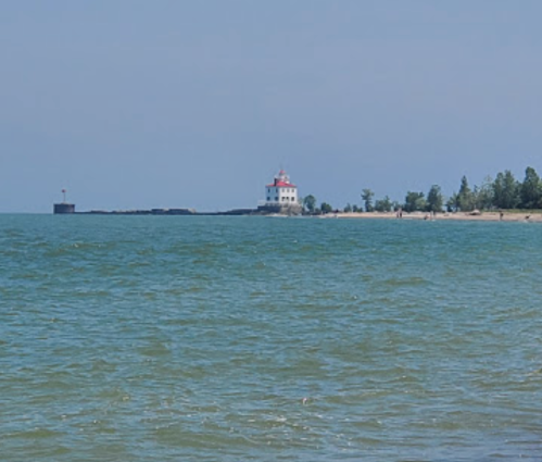 A calm lake scene featuring a lighthouse on the shore, with clear blue water and a sandy beach in the background.