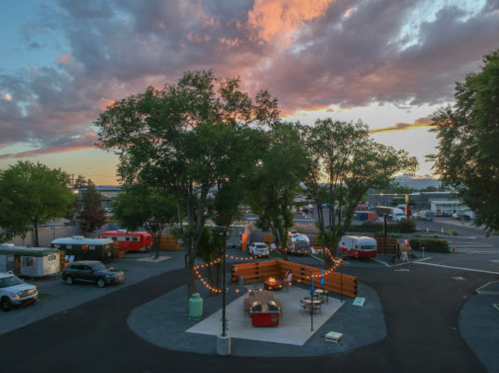 A cozy outdoor space with trees, string lights, and a fire pit, surrounded by parked trailers and a colorful sunset sky.