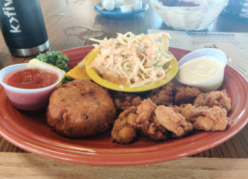 A plate of fried seafood, including crispy fish, a crab cake, coleslaw, and dipping sauces, served with a lemon wedge.