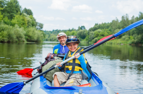 A man and a child smile while kayaking on a calm river, surrounded by lush greenery and a blue sky.