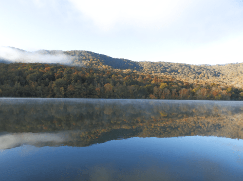 A serene lake reflects a colorful autumn landscape with mist rising from the water and mountains in the background.