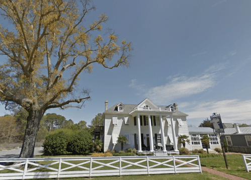 A large white house with a porch, surrounded by trees and a white fence, under a clear blue sky.