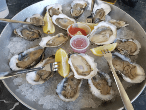 A platter of fresh oysters on ice, garnished with lemon wedges and a small cup of cocktail sauce.