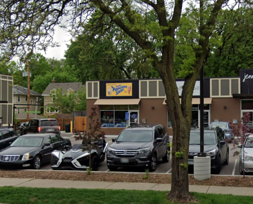 A parking lot with several cars and a storefront featuring a "Popcorn Shop" sign under a tree.