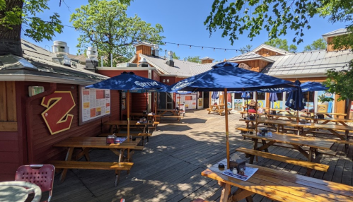 Outdoor dining area with wooden picnic tables, blue umbrellas, and colorful menus on a sunny day.