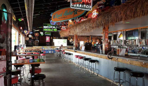 A vibrant bar interior with colorful stools, a thatched roof, and neon signs, featuring a long wooden bar and tables.