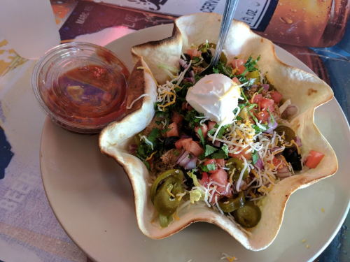 A colorful taco salad in a crispy shell, topped with cheese, sour cream, and fresh vegetables, served with salsa.