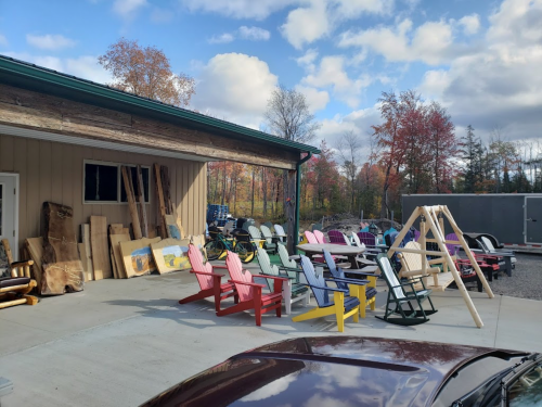 A collection of colorful Adirondack chairs and wooden planks outside a building, surrounded by trees and a cloudy sky.