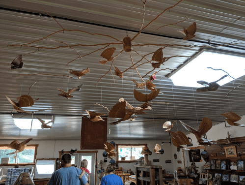 Wooden birds hang from branches in a workshop, with visitors admiring the unique craftsmanship below.
