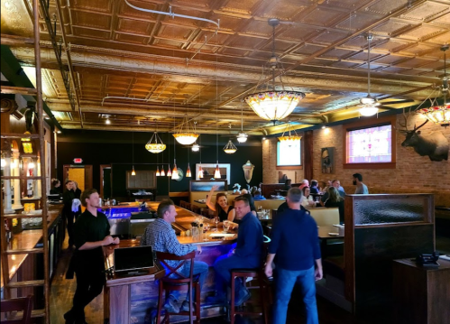 A lively restaurant interior with a bar, patrons dining, and decorative lighting, featuring a vintage ceiling and brick walls.