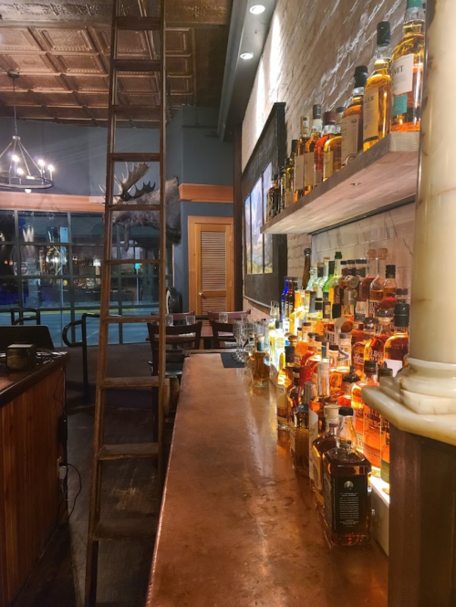 A cozy bar with a polished countertop, shelves lined with various whiskey bottles, and a wooden ladder in the background.