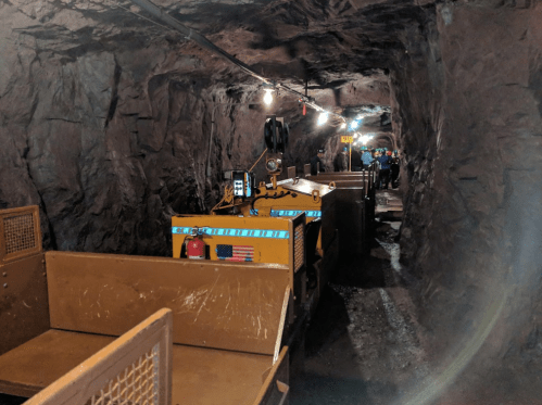 A mining cart in a dimly lit tunnel, with rocky walls and overhead lights illuminating the path ahead.