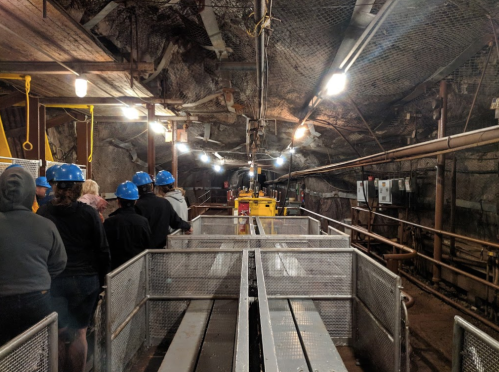 A group of people in blue helmets stands in a dimly lit mine tunnel, with metal walkways and machinery visible.