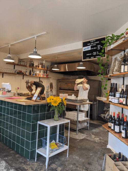 A cozy café interior with a green-tiled counter, a chef preparing food, and shelves stocked with wine and flowers.
